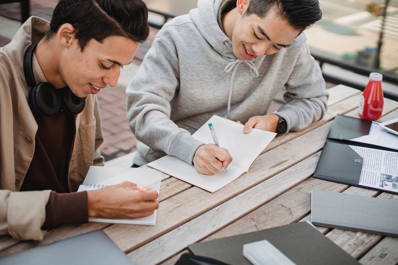 Crop content multiethnic male students in casual clothes taking notes in copybooks while preparing for exam together in campus park